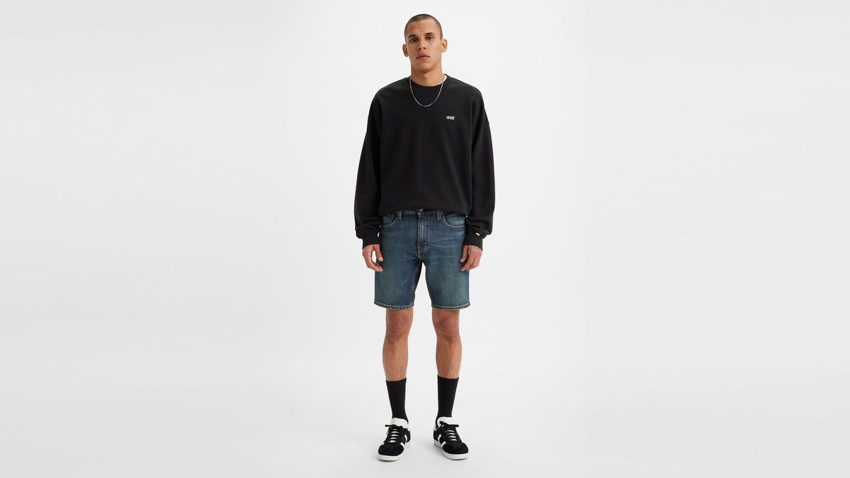 Front view of man in a black sweatshirt & denim shorts, black sneakers, & necklace