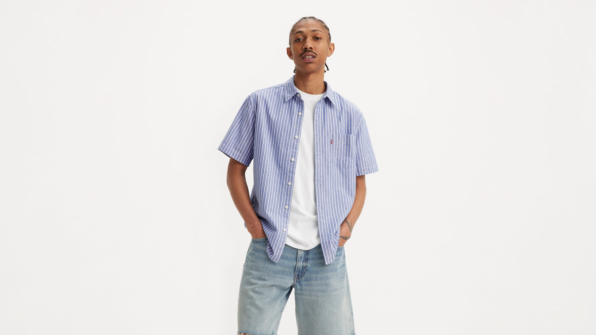 Front view of a man in a striped button-up shirt & denim shorts, hands in pockets