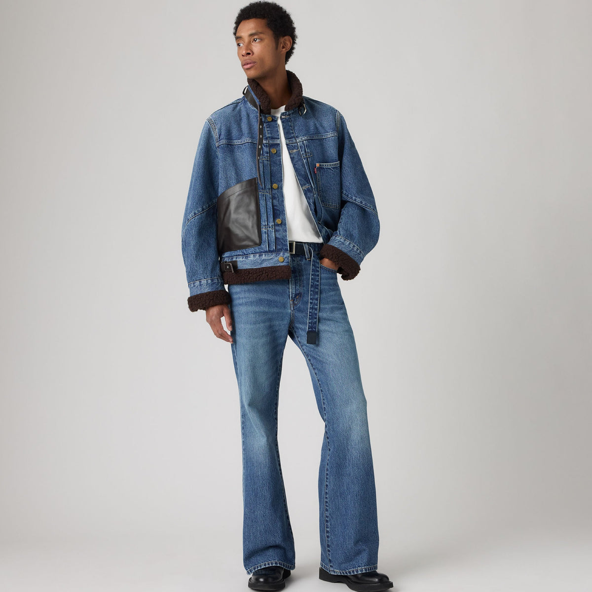 A model wearing a blue denim jacket with brown shearling lining and matching blue denim flare jeans, styled with a white t-shirt and black shoes, against a light gray studio background.